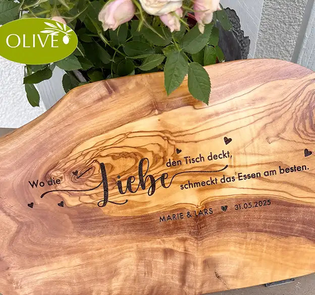 Motiv WO DIE LIEBE - Personalisiertes Schneidebrett aus Olivenholz mit rustikalem Rand und glatter Oberfläche und mit dem text "Wo die Liebe den Tisch deckt, schmeckt das Essen am besten." Darunter die personalisierten Pärchennamen und das Datum. - Jeske Design