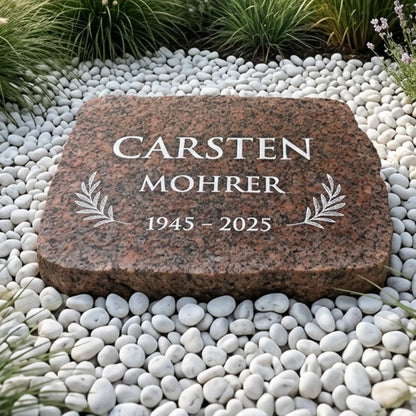 Liegestein aus rotem Granit mit Namen, Lebensdaten und Olivenweigen auf weißem Kiesel