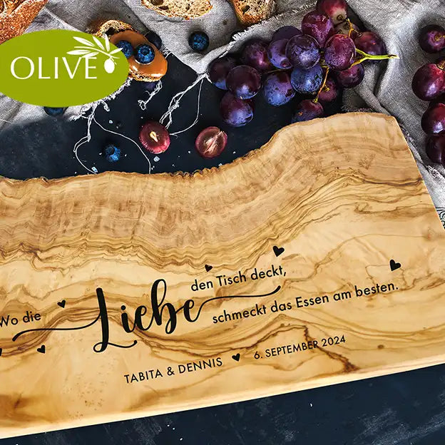 Motiv WO DIE LIEBE - Personalisiertes Schneidebrett aus Olivenholz mit rustikalem Rand und glatter Oberfläche und mit dem text "Wo die Liebe den Tisch deckt, schmeckt das Essen am besten." Darunter die personalisierten Pärchennamen und das Datum. - Jeske Design