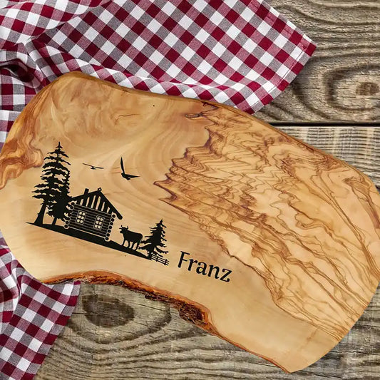 Motiv HÜTTE - Personalisiertes Schneidebrett aus Olivenholz mit rustikalem Rand und glatter Oberfläche mit dem Gravurmotiv einer Holzhütte Almhütte mit Tannen und einer Kuh und zwei Vögel in der Luft. Das Motiv kann personalisiert werden. - Jeske Design