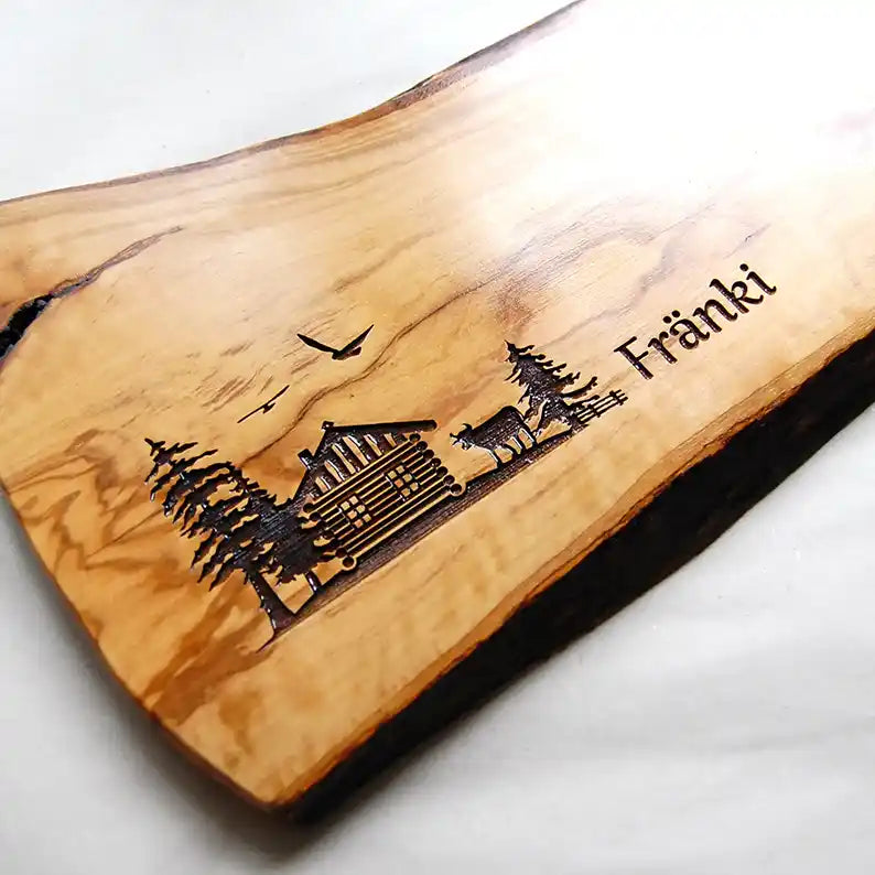 Motiv HÜTTE - Personalisiertes Schneidebrett aus Olivenholz mit rustikalem Rand und glatter Oberfläche mit dem Gravurmotiv einer Holzhütte Almhütte mit Tannen und einer Kuh und zwei Vögel in der Luft. Das Motiv kann personalisiert werden. - Jeske Design