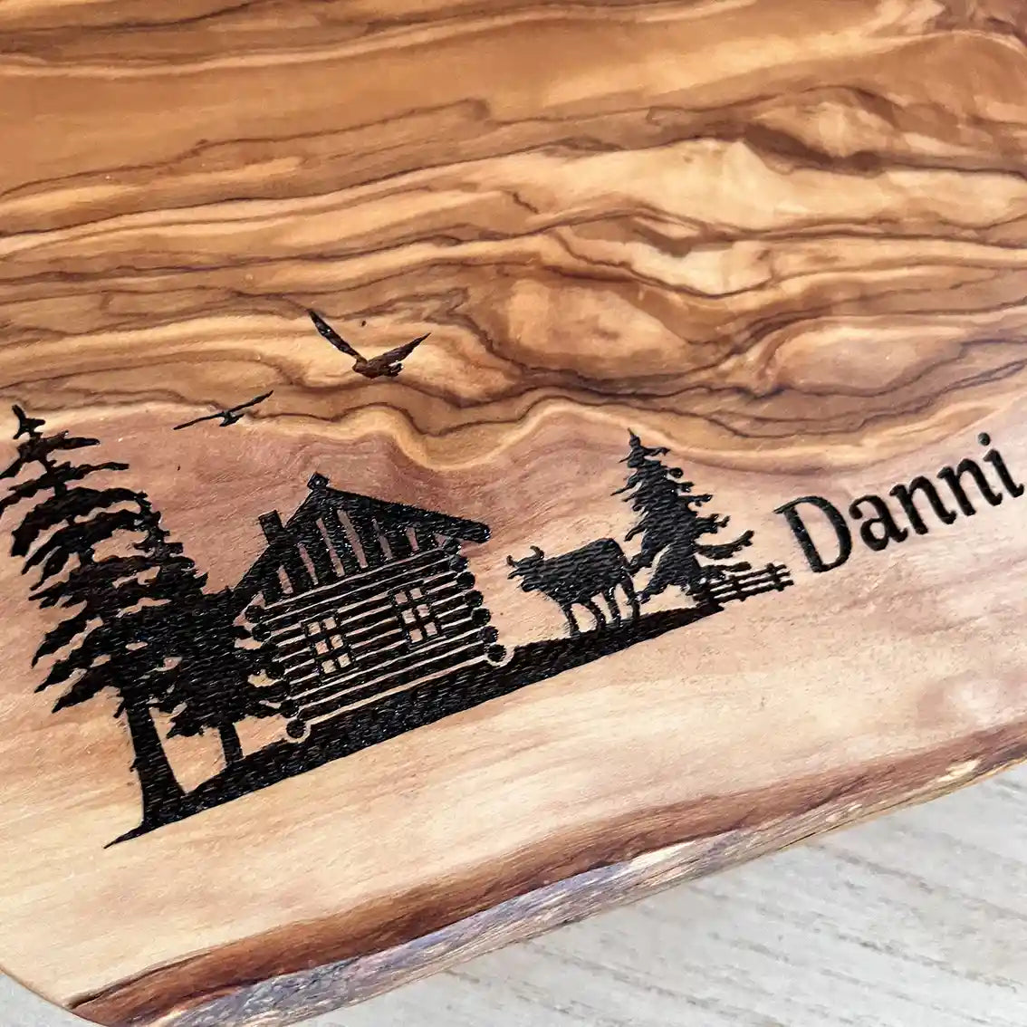 Motiv HÜTTE - Personalisiertes Schneidebrett aus Olivenholz mit rustikalem Rand und glatter Oberfläche mit dem Gravurmotiv einer Holzhütte Almhütte mit Tannen und einer Kuh und zwei Vögel in der Luft. Das Motiv kann personalisiert werden. - Jeske Design