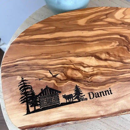 Motiv HÜTTE - Personalisiertes Schneidebrett aus Olivenholz mit rustikalem Rand und glatter Oberfläche mit dem Gravurmotiv einer Holzhütte Almhütte mit Tannen und einer Kuh und zwei Vögel in der Luft. Das Motiv kann personalisiert werden. - Jeske Design