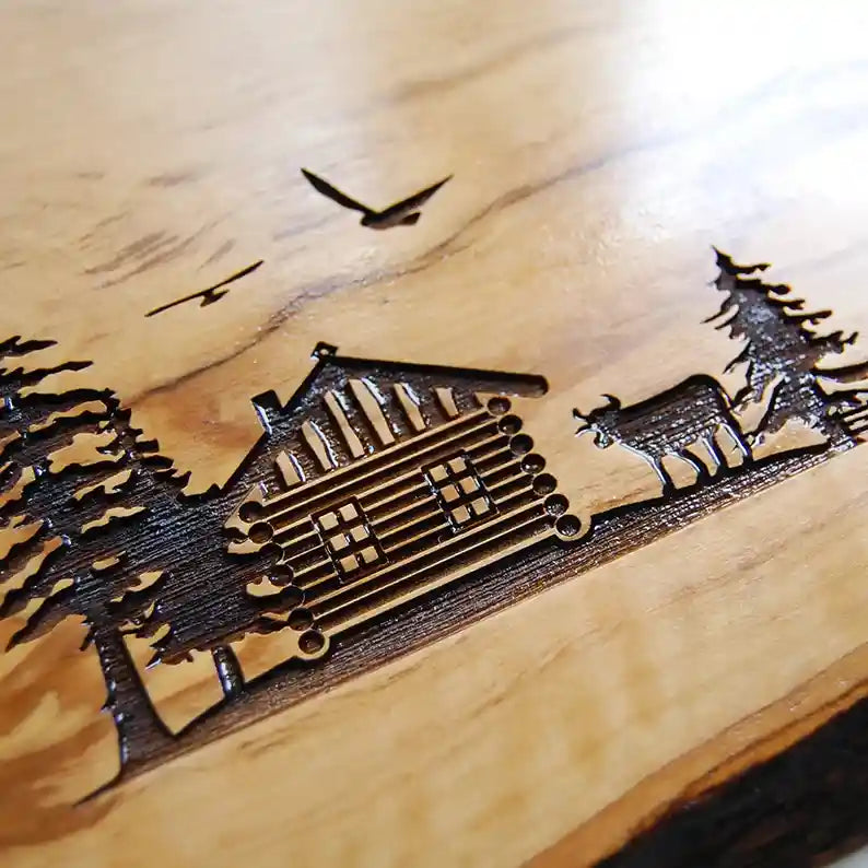 Motiv HÜTTE - Personalisiertes Schneidebrett aus Olivenholz mit rustikalem Rand und glatter Oberfläche mit dem Gravurmotiv einer Holzhütte Almhütte mit Tannen und einer Kuh und zwei Vögel in der Luft. Das Motiv kann personalisiert werden. - Jeske Design