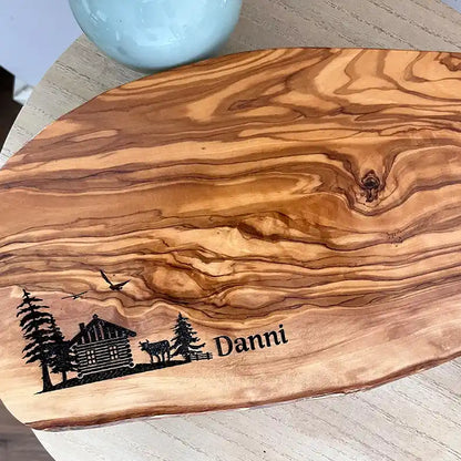 Motiv HÜTTE - Personalisiertes Schneidebrett aus Olivenholz mit rustikalem Rand und glatter Oberfläche mit dem Gravurmotiv einer Holzhütte Almhütte mit Tannen und einer Kuh und zwei Vögel in der Luft. Das Motiv kann personalisiert werden. - Jeske Design
