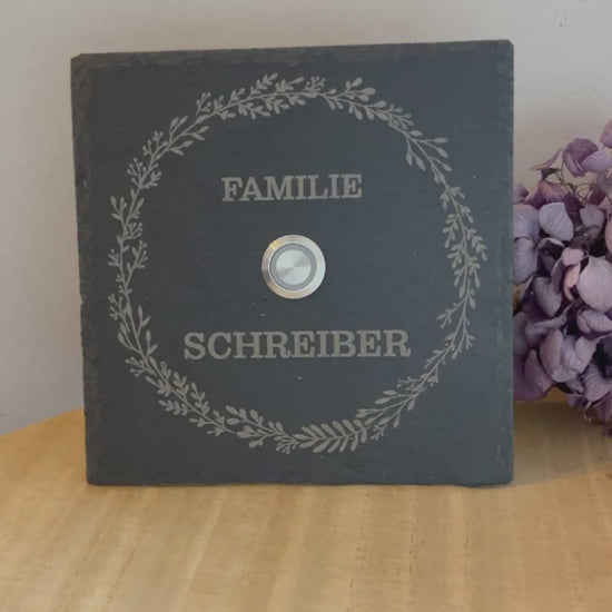 Personalisiertes Klingelschild aus Schiefer mit Edelstahl Taster mit LEDBeleuchtung oder ohne Beleuchtung. Zarter Blätterkranz rund mit Klingelknopf innen. Text in klassischer Druckschrift 2-zeilig oben und unten in der Größe ø 15 cm rund oder 15x15 cm, 20x15 cm, 26x16 cm oder 30x20 cm.