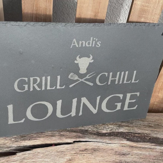 Dekoschild aus Schiefer mit dem Motiv Grill & Chill Lounge, welches auf eine Schieferplatte in der Größe 30 x 20 cm  und personalisierter Form mittels Laser graviert wird. Zwischen Grill & Chill sind gekreuzte Grillgabeln und ein stilisierter Bullenkopf.