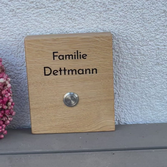 Hochwertiges Klingelschild aus Eiche in der Größe 12 x 14 cm mit moderner schlichte Schrift zweizeilig oberhalb des Tasters mit Edelstahltaster und 2 Kunststoffhalter auf der Rückseite zum Befestigen an die Hauswand