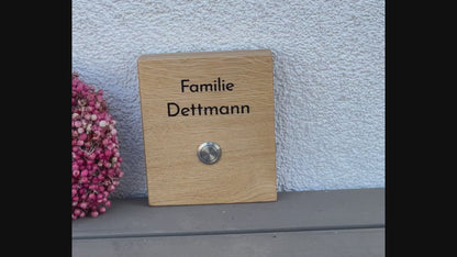 Hochwertiges Klingelschild aus Eiche in der Größe 12 x 14 cm mit moderner schlichte Schrift zweizeilig oberhalb des Tasters mit Edelstahltaster und 2 Kunststoffhalter auf der Rückseite zum Befestigen an die Hauswand
