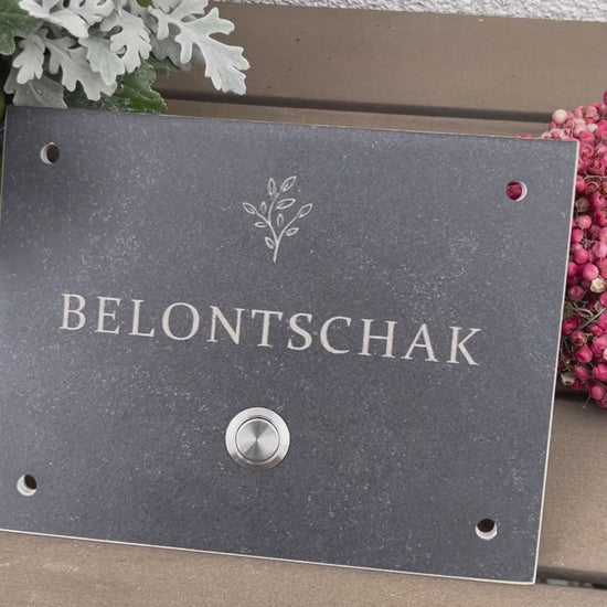 Dunkelgrau-meliertes Klingelschild aus Feinsteinzeug-Keramik frostsicher in der Größe 20 x 15 cm mit dem Wunschnamen und dem Motiv einer stilisierten Pflanze graviert. Verschiedene Taster zur Auswahl: Edelstahl matt (ohne/mit LED), Edelstahl goldfarben, Aluminium schwarz. Hier im mit Edelstahltaster matt ohne LED und 4-Loch-Ecke für die Befestigung an die Hauswand