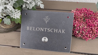 Dunkelgrau-meliertes Klingelschild aus Feinsteinzeug-Keramik frostsicher in der Größe 20 x 15 cm mit dem Wunschnamen und dem Motiv einer stilisierten Pflanze graviert. Verschiedene Taster zur Auswahl: Edelstahl matt (ohne/mit LED), Edelstahl goldfarben, Aluminium schwarz. Hier im mit Edelstahltaster matt ohne LED und 4-Loch-Ecke für die Befestigung an die Hauswand