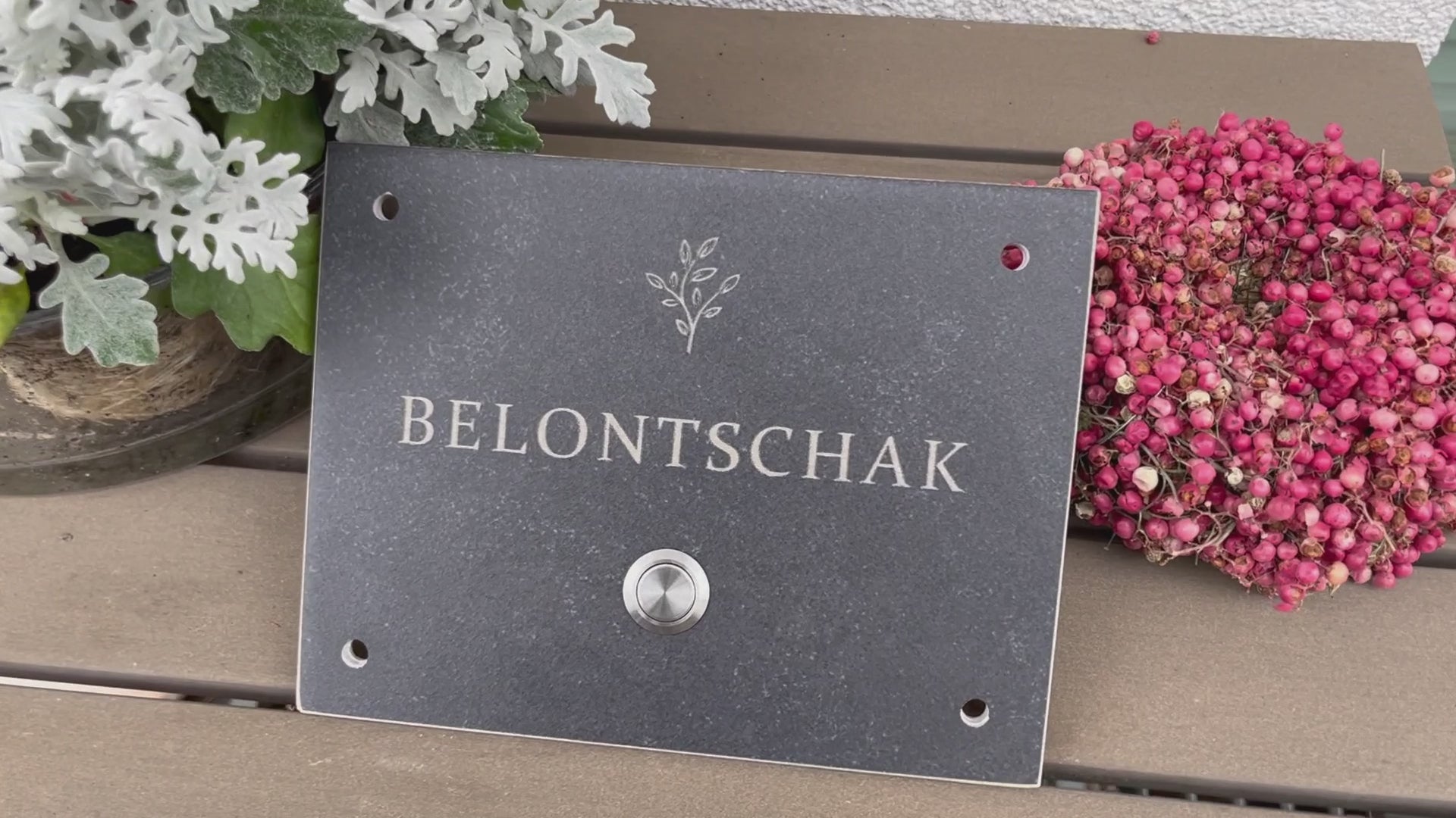 Dunkelgrau-meliertes Klingelschild aus Feinsteinzeug-Keramik frostsicher in der Größe 20 x 15 cm mit dem Wunschnamen und dem Motiv einer stilisierten Pflanze graviert. Verschiedene Taster zur Auswahl: Edelstahl matt (ohne/mit LED), Edelstahl goldfarben, Aluminium schwarz. Hier im mit Edelstahltaster matt ohne LED und 4-Loch-Ecke für die Befestigung an die Hauswand