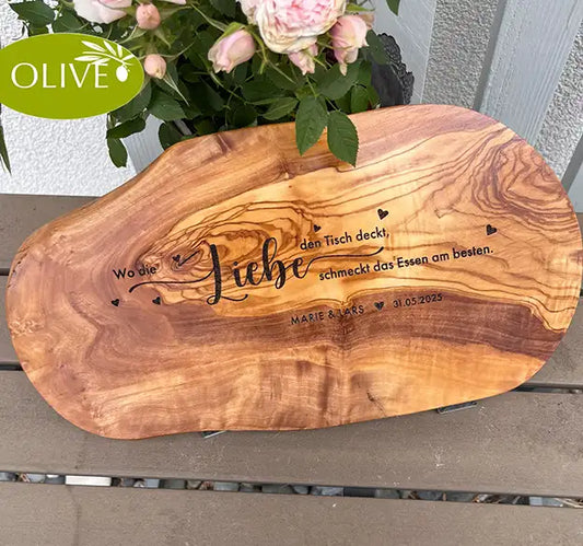 Motiv WO DIE LIEBE - Personalisiertes Schneidebrett aus Olivenholz mit rustikalem Rand und glatter Oberfläche und mit dem text "Wo die Liebe den Tisch deckt, schmeckt das Essen am besten." Darunter die personalisierten Pärchennamen und das Datum. - Jeske Design